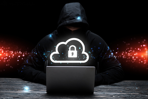 hacker v kapuci pomocí notebooku v blízkosti cloudu s visacím zámkem na černé  - Fotografie, Obrázek