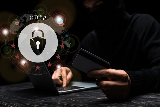 selective focus of hacker in mask using laptop while holding credit card near padlock with gdpr lettering on black  - Foto, imagen