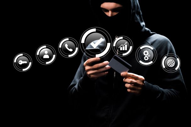 hacker in kap met behulp van smartphone en het houden van credit card in de buurt van web iconen op zwart  - Foto, afbeelding