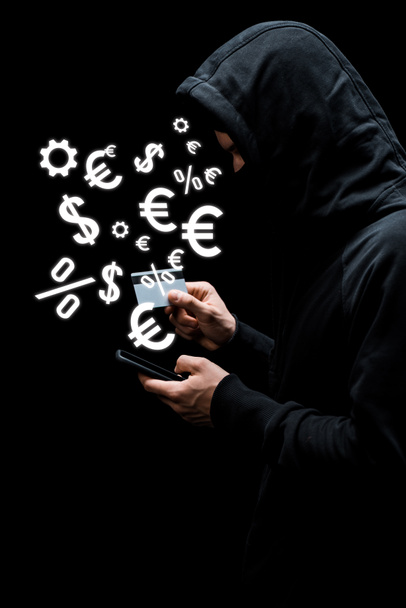 hacker en la capucha y la máscara con el teléfono inteligente y la celebración de tarjetas de crédito cerca de signos de dinero aislados en negro
  - Foto, Imagen