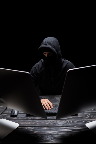 κουκουλοφόρος άνδρας με μάσκα κάθεται κοντά σε οθόνες υπολογιστών απομονωμένες σε μαύρο  - Φωτογραφία, εικόνα