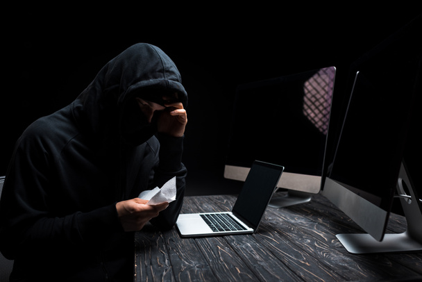 upset hacker holding tissue near laptop and computer monitors with blank screen isolated on black  - Fotó, kép