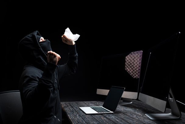 emotional hacker holding tissue near laptop and computer monitors with blank screen isolated on black  - Photo, Image