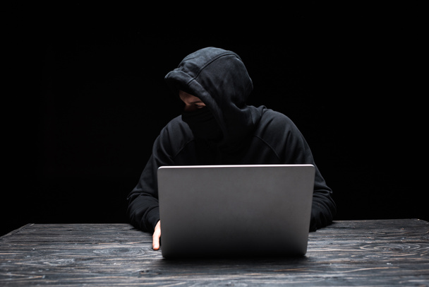 junger Hacker in Maske schaut weg, während er Laptop isoliert auf schwarz benutzt  - Foto, Bild