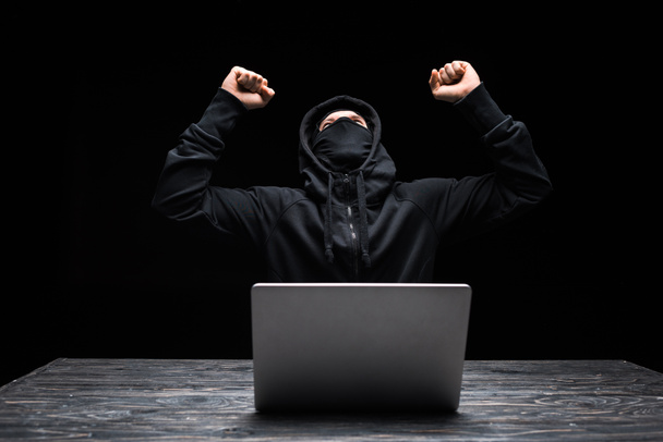 podekscytowany haker w masce świętując triumf w pobliżu laptopa odizolowany na czarno  - Zdjęcie, obraz