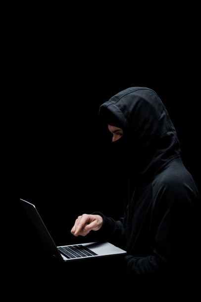 hacker in hood and mask pointing with finger at laptop isolated on black  - Photo, Image