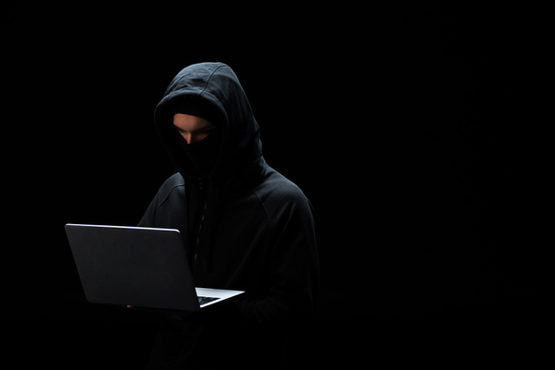 hacker en máscara y capucha usando portátil aislado en negro
  - Foto, imagen