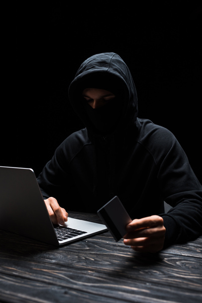 hacker in hood using laptop while holding credit card isolated on black  - Φωτογραφία, εικόνα