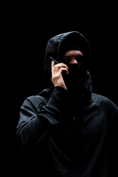 hacker in hood and mask talking on smartphone isolated on black  - Photo, Image