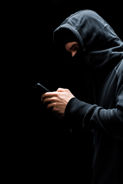 anonieme hacker in kap en masker met behulp van smartphone geïsoleerd op zwart  - Foto, afbeelding