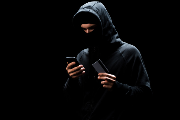 hacker in mask using smartphone and holding credit card isolated on black  - Photo, Image
