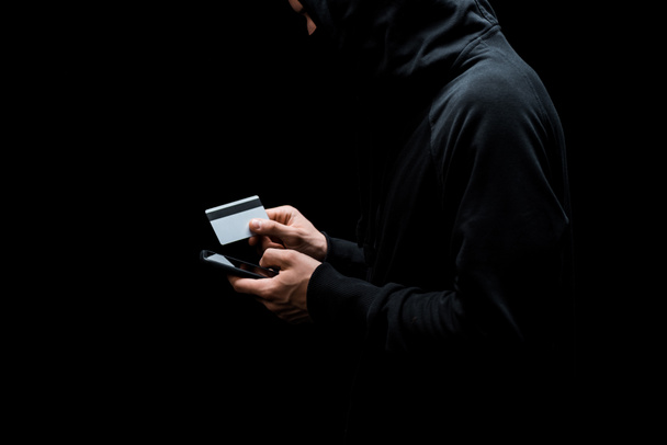 cropped view of hacker in hood and mask using smartphone and holding credit card isolated on black  - Photo, Image