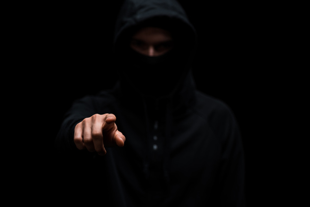 enfoque selectivo de hacker en la capucha y máscara apuntando con el dedo aislado en negro
  - Foto, imagen
