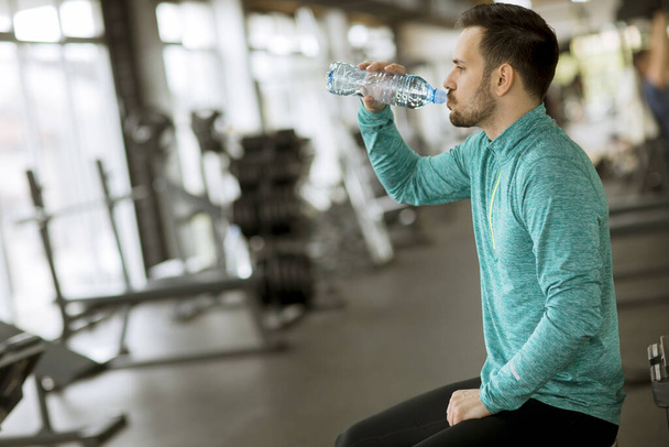 Handsome young sportsman drinking water from  bottle in the gym and resting - Photo, Image