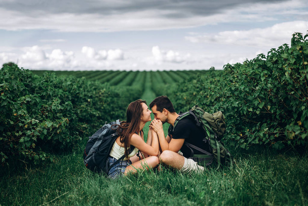 Man and woman with backpacks on their backs sitting hugging on the green grass between the currant bushes. Walking on the currant plantations in the countryside - Fotografie, Obrázek