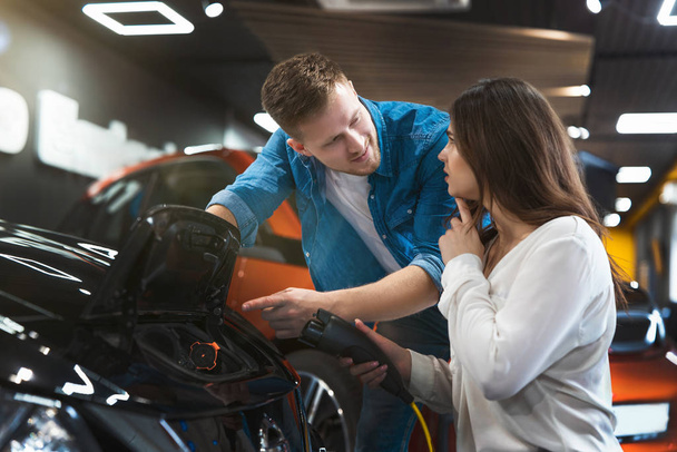 handsome man shows beautiful brunette woman how to use charging cable for electric car sitting near open bonnet , looking happy and excited, eco friendly concept - Φωτογραφία, εικόνα