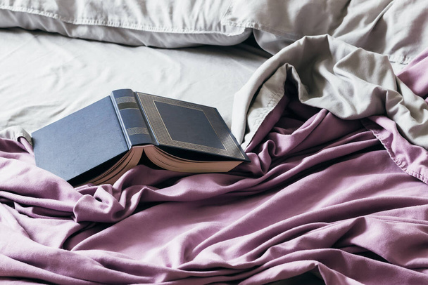 Ανοιχτό μπλε βιβλίο στο κρεβάτι με μωβ κάλυμμα κρεβατιού και γκρι μαξιλάρια. Συναρπαστικό βιβλίο σε μαλακό κρεβάτι - Φωτογραφία, εικόνα