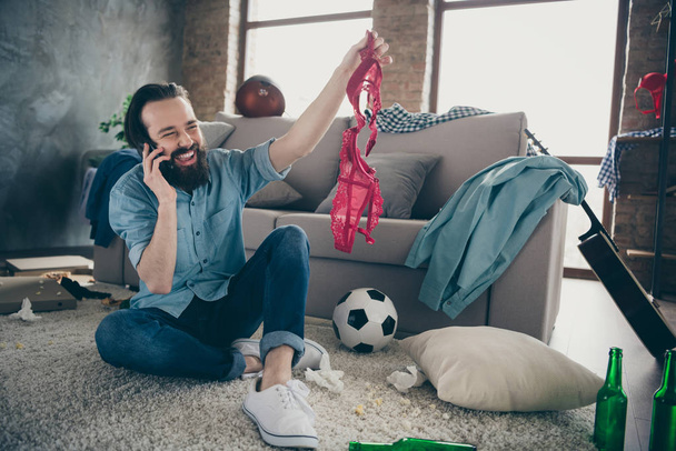 Photo of laughing hipster guy holding telephone telling friends intimacy details of his active naughty life night bad person watch red bra hands sitting floor trash after party flat indoors - Foto, Imagem