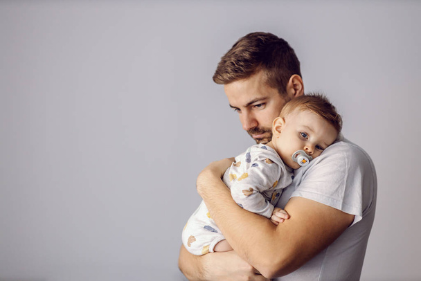 Young good-looking unshaven caring father holding his beloved only son and hugging him. Adorable little boy leaning on father's shoulder, having pacifier in mouth. It's noon nap time - Photo, Image