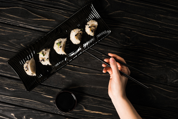 partial view of woman eating delicious Chinese boiled dumpling with chopsticks and soy sauce at black wooden table - Foto, immagini