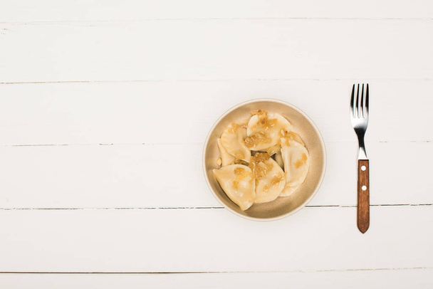 τοπ άποψη του νόστιμα varenyky με τηγανητό κρεμμύδι σε μπολ κοντά πιρούνι σε λευκό ξύλινο τραπέζι - Φωτογραφία, εικόνα