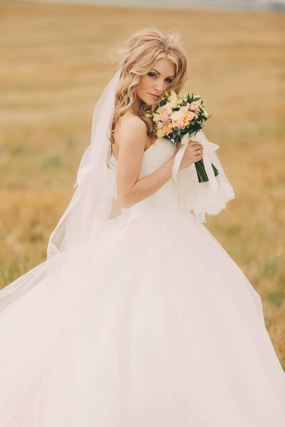 バラの花束と豪華な白いウェディングドレスの美しい若い花嫁。夏のフィールドでかわいい花嫁の肖像画。結婚式の日だメイクや髪型と美しい花嫁. - 写真・画像