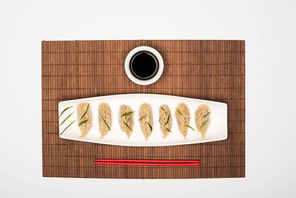 vrchní pohled na lahodné čínské vařené knedlíky na talíři v blízkosti hůlek a sójové omáčky na bambusové rohoži na bílém pozadí - Fotografie, Obrázek