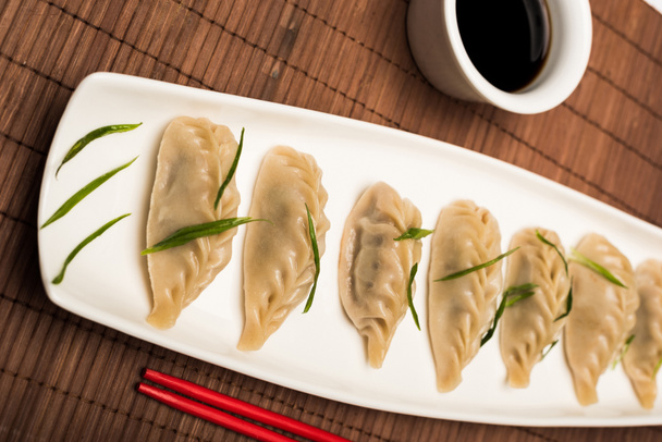 heerlijke Chinese gekookte knoedels op bord bij eetstokjes en sojasaus op bamboe mat - Foto, afbeelding