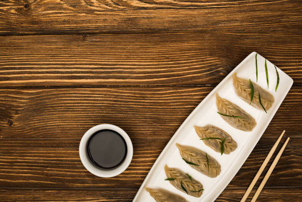 bovenaanzicht van heerlijke Chinese gekookte knoedels op bord bij eetstokjes en sojasaus op houten tafel - Foto, afbeelding