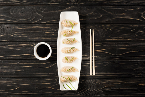 bovenaanzicht van heerlijke Chinese gekookte knoedels op bord bij eetstokjes en sojasaus op zwarte houten tafel - Foto, afbeelding