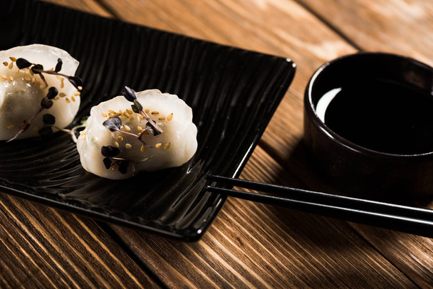 close-up zicht op heerlijke Chinese gekookte knoedels op bord bij eetstokjes en sojasaus op houten tafel - Foto, afbeelding