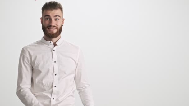 A smiling young bearded man wearing a white shirt is pointing to the side with both hands and open mouth isolated over white wall background - Záběry, video