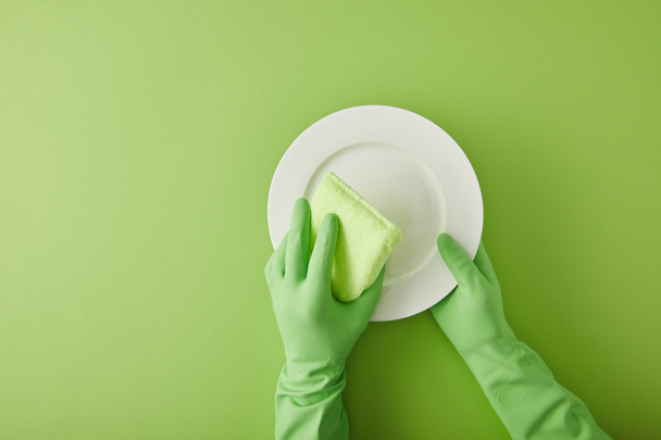 Lastik eldivenli temizlikçi yeşil üzerinde süngerle tabağı yıkıyor.  - Fotoğraf, Görsel