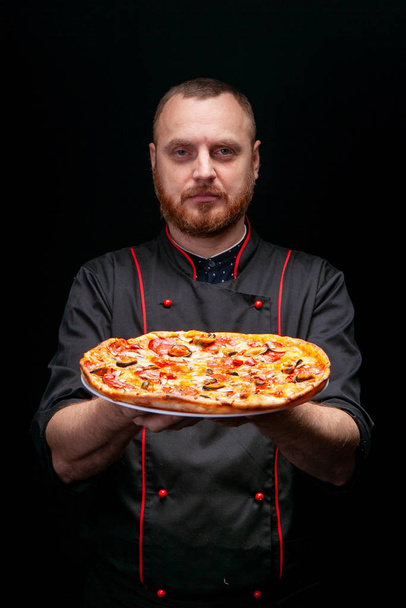 mannelijke kok in een zwarte tuniek en handschoenen houdt een verse pizza met olijven voor zich - Foto, afbeelding