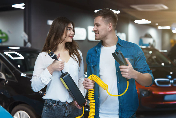young family couple handsome man and beautiful brunette woman holding charging cable for electric car , looking happy - Foto, imagen