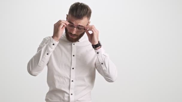 A calm young bearded man wearing a white shirt takes off his glasses and touches his bridge of nose isolated over white wall background - Filmati, video