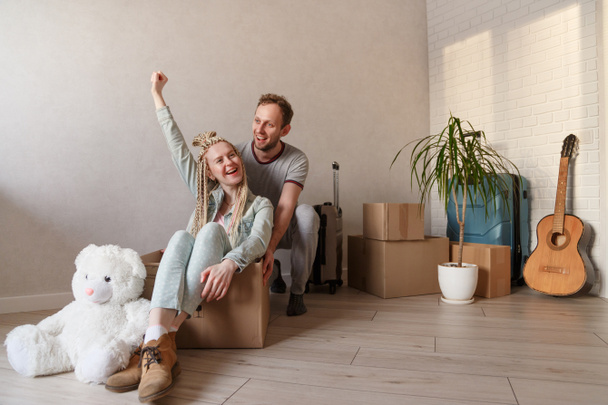 幸せな若いカップルは新しいアパートに移動します。男は腕を上げて箱の中に座っている女性を押し出す。背景に詰め込まれたもの. - 写真・画像