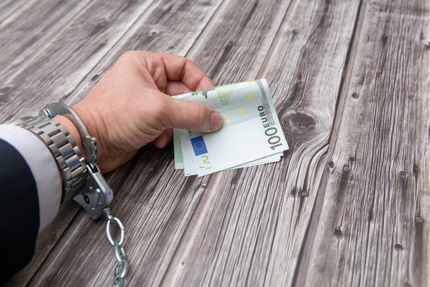 Een man 's hand in handboeien en ons dollars bankbiljetten op houten tafel. Concept van corruptie of economische criminaliteit. - Foto, afbeelding