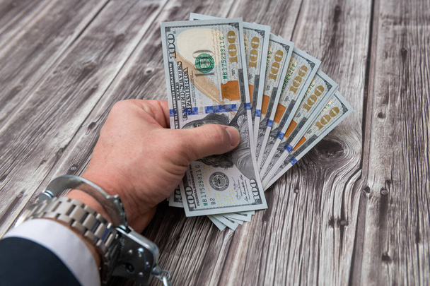 Een man 's hand in handboeien en ons dollars bankbiljetten op houten tafel. Concept van corruptie of economische criminaliteit. - Foto, afbeelding