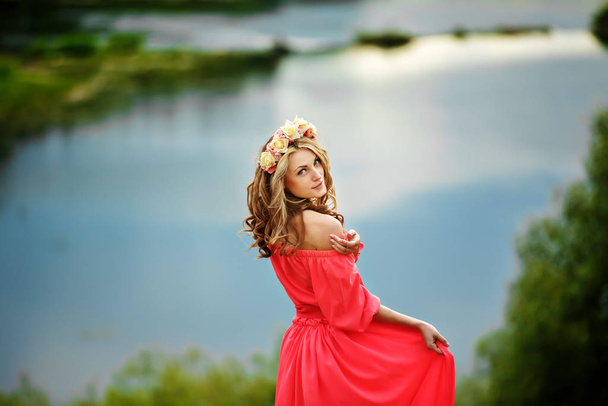 beautiful young blonde woman with a flower wreath on her head. Beauty girl with flowers hairstyle in a red long dress near the lake. Fashion photo, copy space - Photo, Image