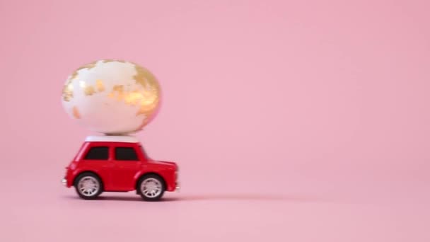 Little red toy car with a golden egg on the hood on a pink background. Gift delivery. Happy easter concept - Footage, Video