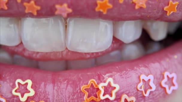 Vista macro de labios femeninos con estrellas doradas decorativas que muestran la lengua entre dientes blancos perfectos
 - Metraje, vídeo