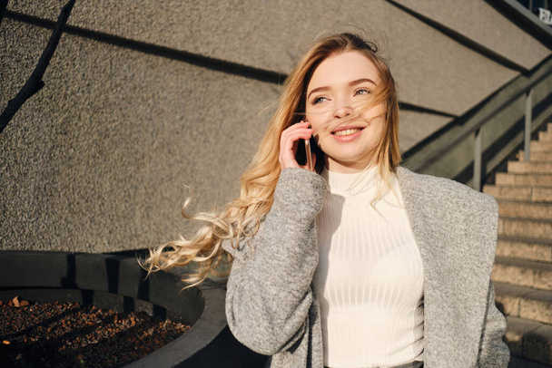Молодая красивая блондинка разговаривает по мобильному телефону счастливо гуляя по улице в солнечный день
 - Фото, изображение