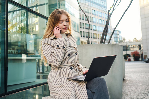 Молодая стильная деловая женщина с ноутбуком уверенно разговаривает по мобильному телефону, работая на городской улице в одиночку
 - Фото, изображение