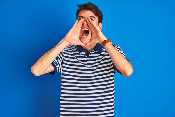Tini fiú visel alkalmi póló áll a kék elszigetelt háttér Kiabálás dühös ki hangosan a kezét a száját - Fotó, kép