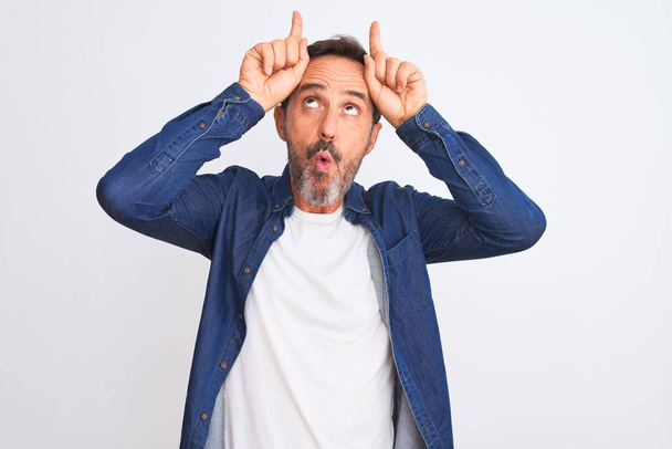 Middle age handsome man wearing blue denim shirt standing over isolated white background doing funny gesture with finger over head as bull horns - Photo, Image