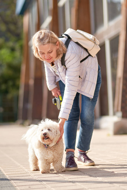 Mulher loira adulta andando com cachorro branco fofo na cidade de verão
 - Foto, Imagem