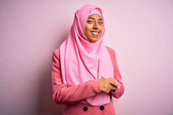 Joven afroamericana afro mujer con hiyab musulmán sobre fondo rosa aislado con las manos juntas y los dedos cruzados sonriendo relajada y alegre. Éxito y optimismo
 - Foto, Imagen