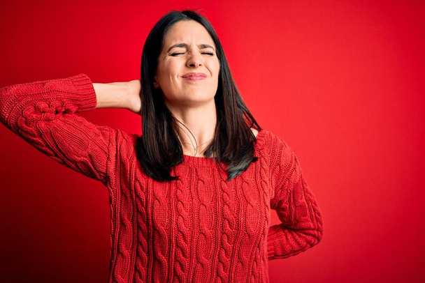 Young brunette woman with blue eyes wearing casual sweater over isolated red background Suffering of neck ache injury, touching neck with hand, muscular pain - Fotografie, Obrázek