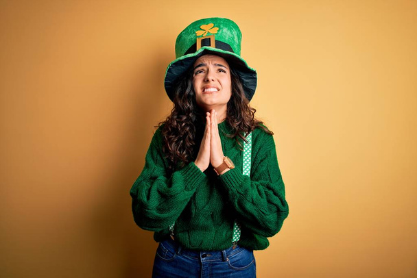 schöne Frau mit lockigem Haar trägt grünen Hut mit Klee feiert den Heiligen Patricks Tag betteln und beten mit den Händen zusammen mit Hoffnung Ausdruck im Gesicht sehr emotional und besorgt. Betteln. - Foto, Bild
